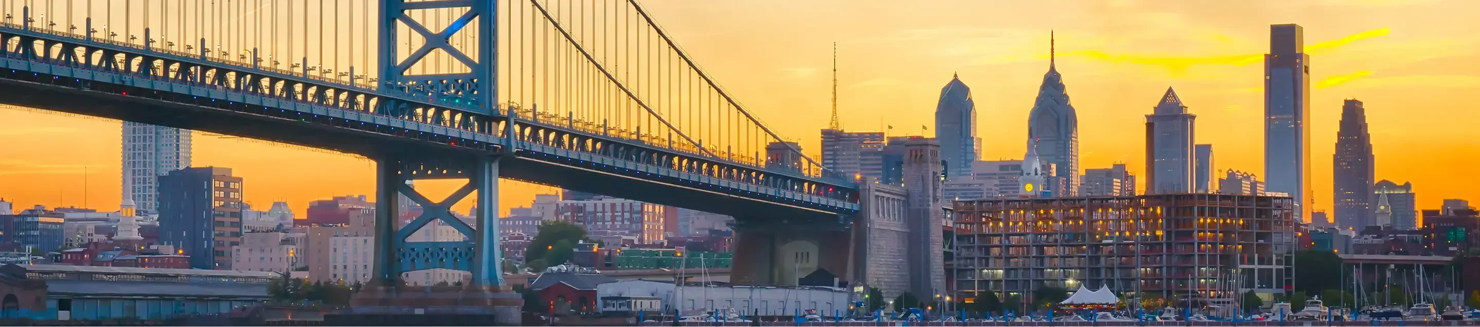 A sunset view of the Benjamin Franklin Bridge with Philadelphia's skyline in the background. Warm colors illuminate the cityscape.