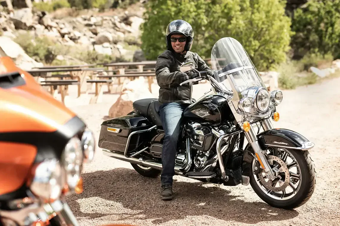 A rider in a leather jacket sits on a sleek black motorcycle, surrounded by greenery and rocky terrain. A vibrant orange bike is nearby.
