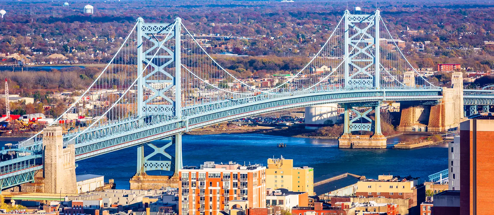 Aerial view of a blue suspension bridge spanning a river, surrounded by a cityscape and autumn foliage in the background.
